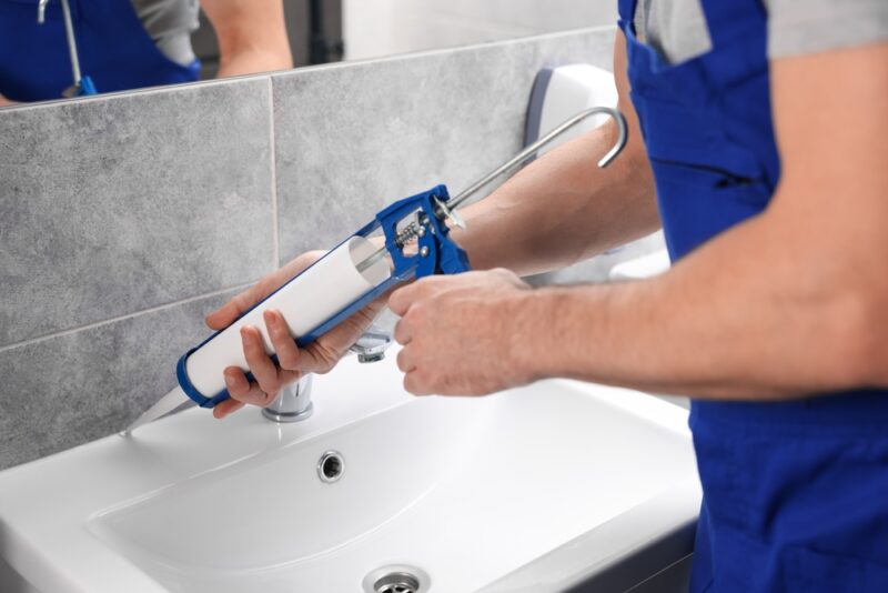 Nanošenje novog sanitarnog silikona oko umivaonika u kupatilu pomoću pištolja za silikon tokom redovnog održavanja.